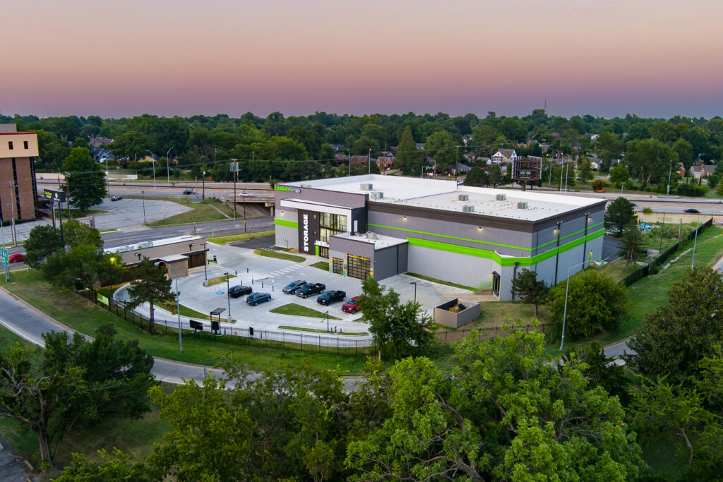 Aerial view of a modern, gray and green climate-controlled storage facility with several cars parked outside, surrounded by trees and a suburban neighborhood at sunset.