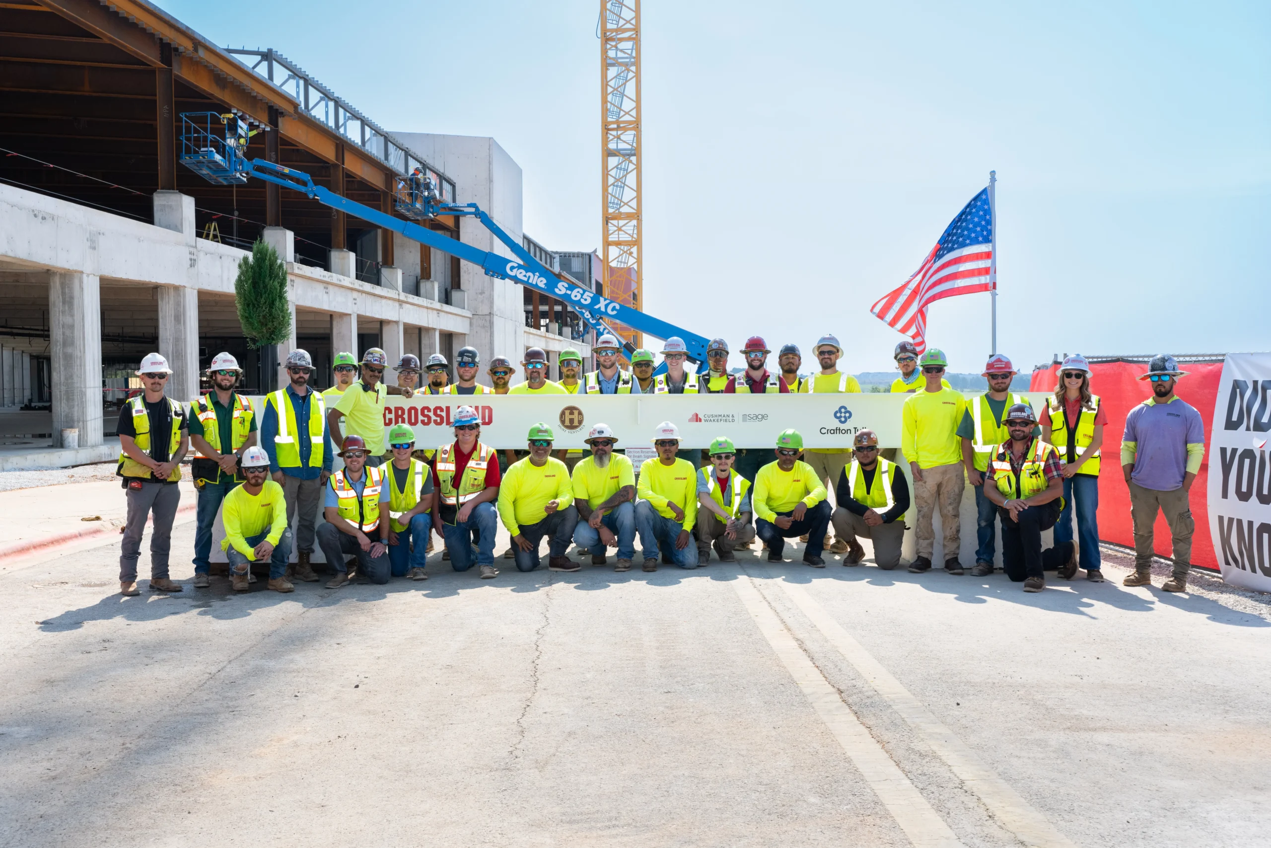 A large group of construction workers in safety gear pose for a group photo in front of an unfinished building, holding a banner, with an American flag and a crane in the background.