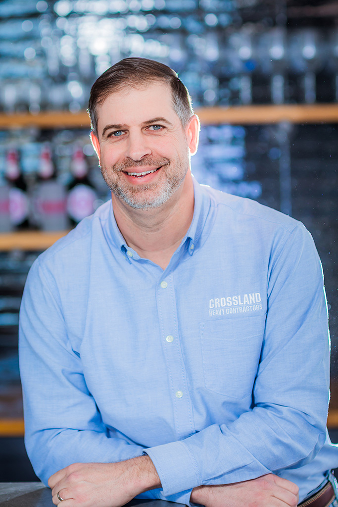 A man with short hair and a beard, wearing a light blue “Crossland Heavy Contractors” shirt, smiles while sitting indoors with blurred bottles and shelves in the background.