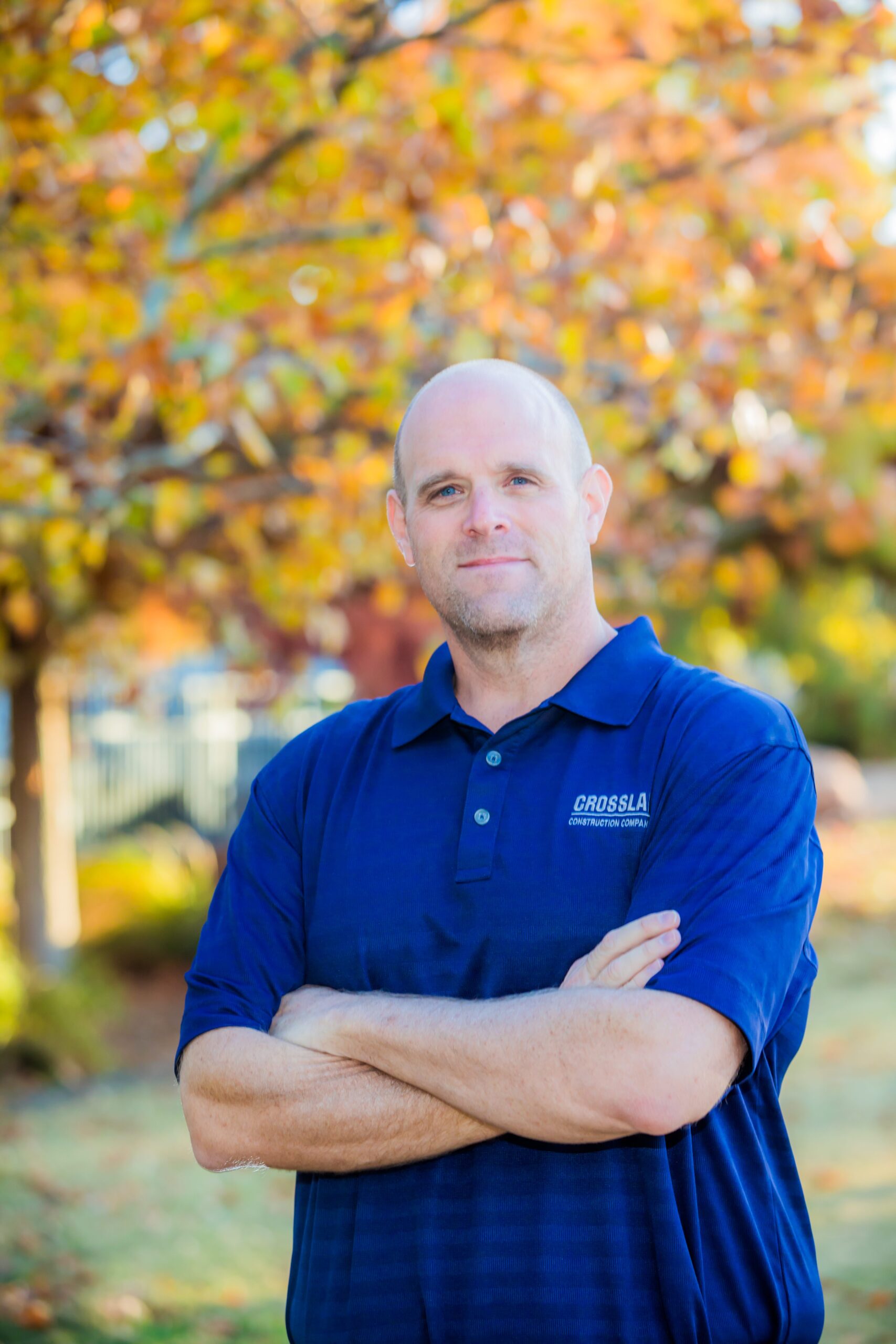 A man with a shaved head stands outdoors in front of autumn trees, wearing a blue collared shirt with "CROSSLA" embroidered on it, arms crossed, looking at the camera with a neutral expression.
