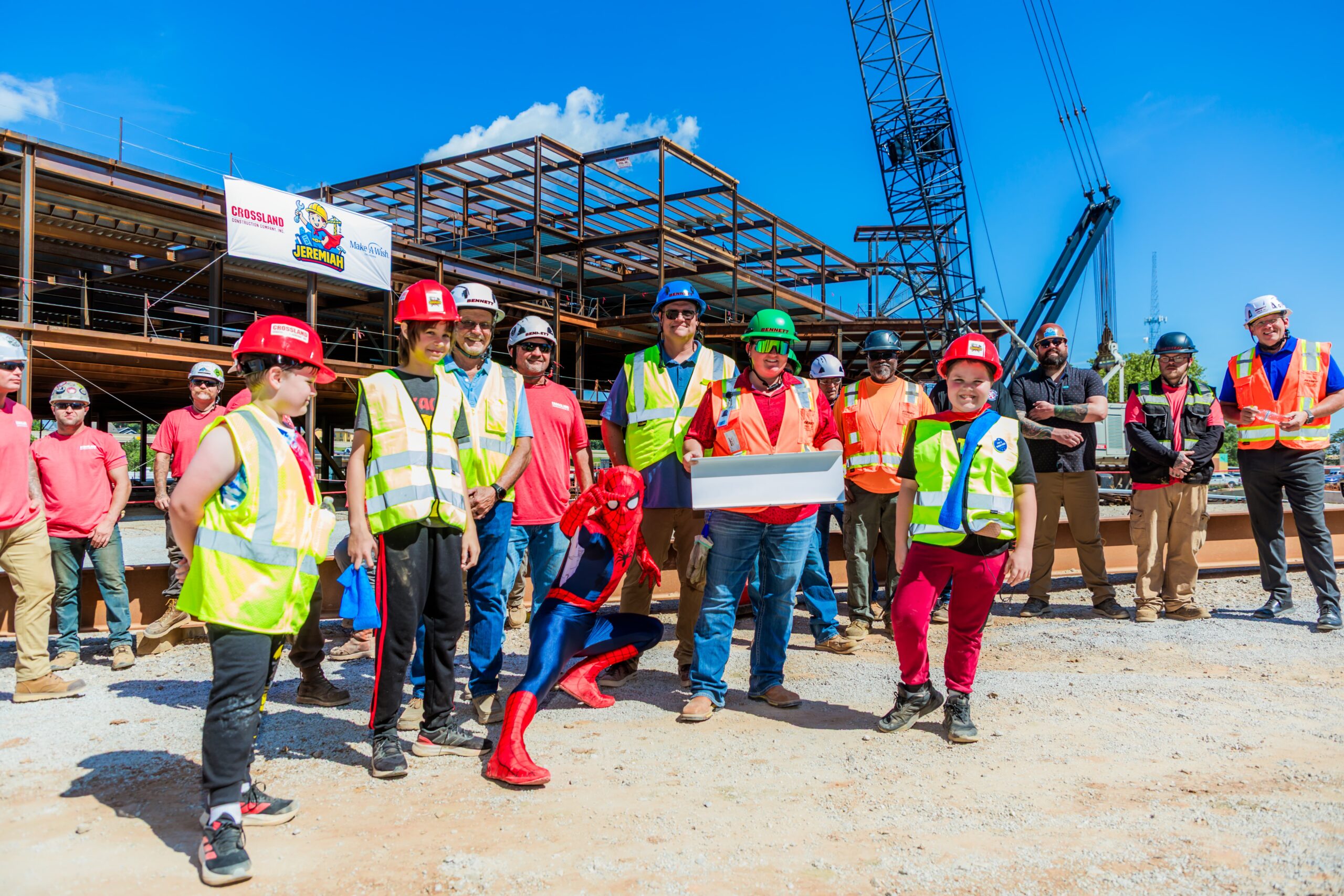 A group of construction workers in safety gear pose at a building site with steel framework; a person dressed as Spider-Man crouches in front, and one worker holds a white box. A crane and blue sky are in the background.