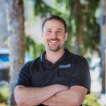A man with short brown hair and a beard smiles with his arms crossed, standing outdoors in front of trees. He wears a black polo shirt with a small “CRISSLANE CONSTRUCTION” logo on the chest.