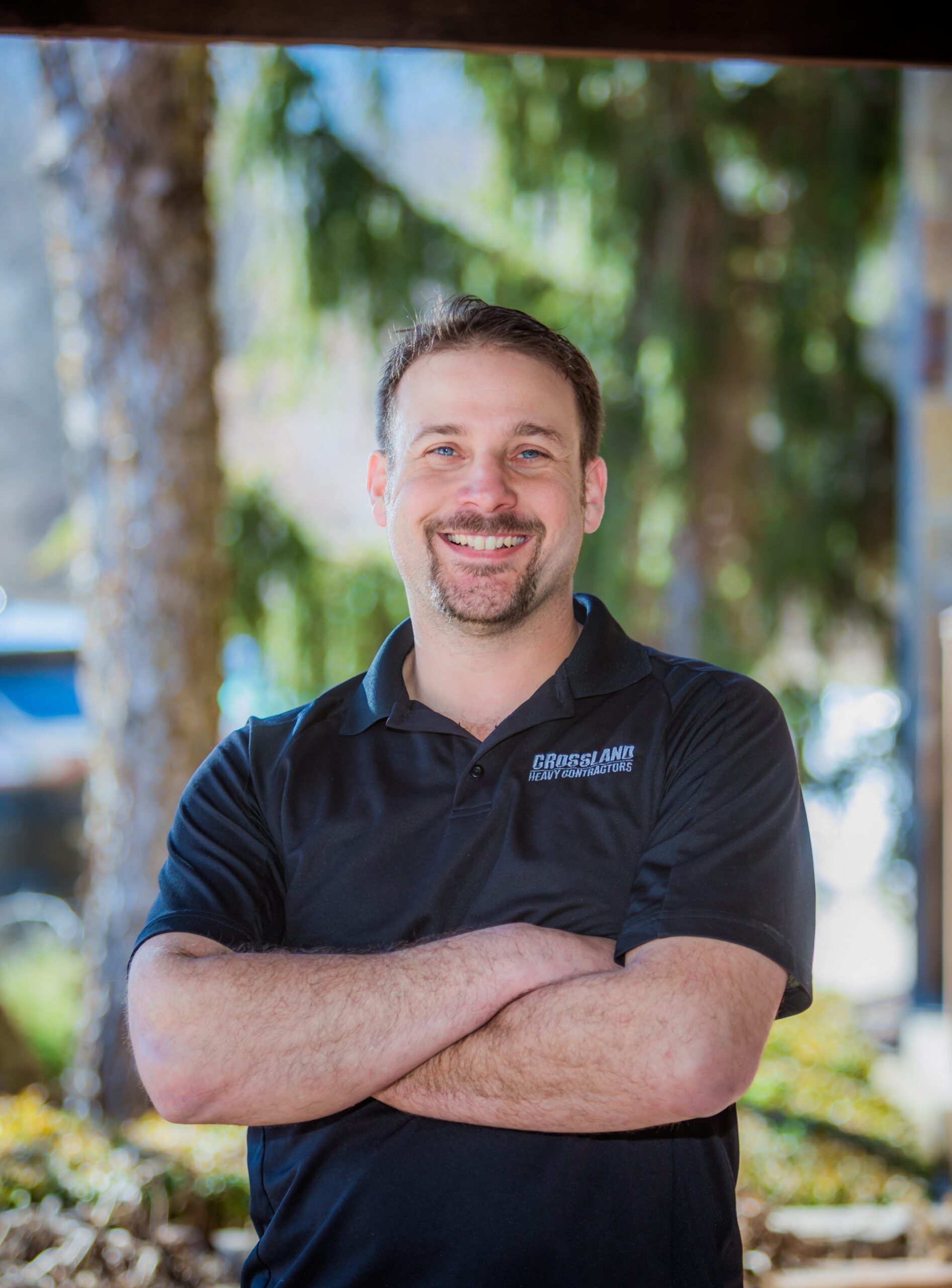 A man with short brown hair and a beard smiles with his arms crossed, standing outdoors in front of trees. He wears a black polo shirt with a small “CRISSLANE CONSTRUCTION” logo on the chest.
