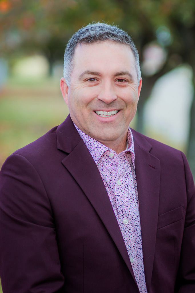A man with short gray hair smiles at the camera, wearing a maroon blazer and a patterned shirt, standing outdoors with blurred trees in the background.