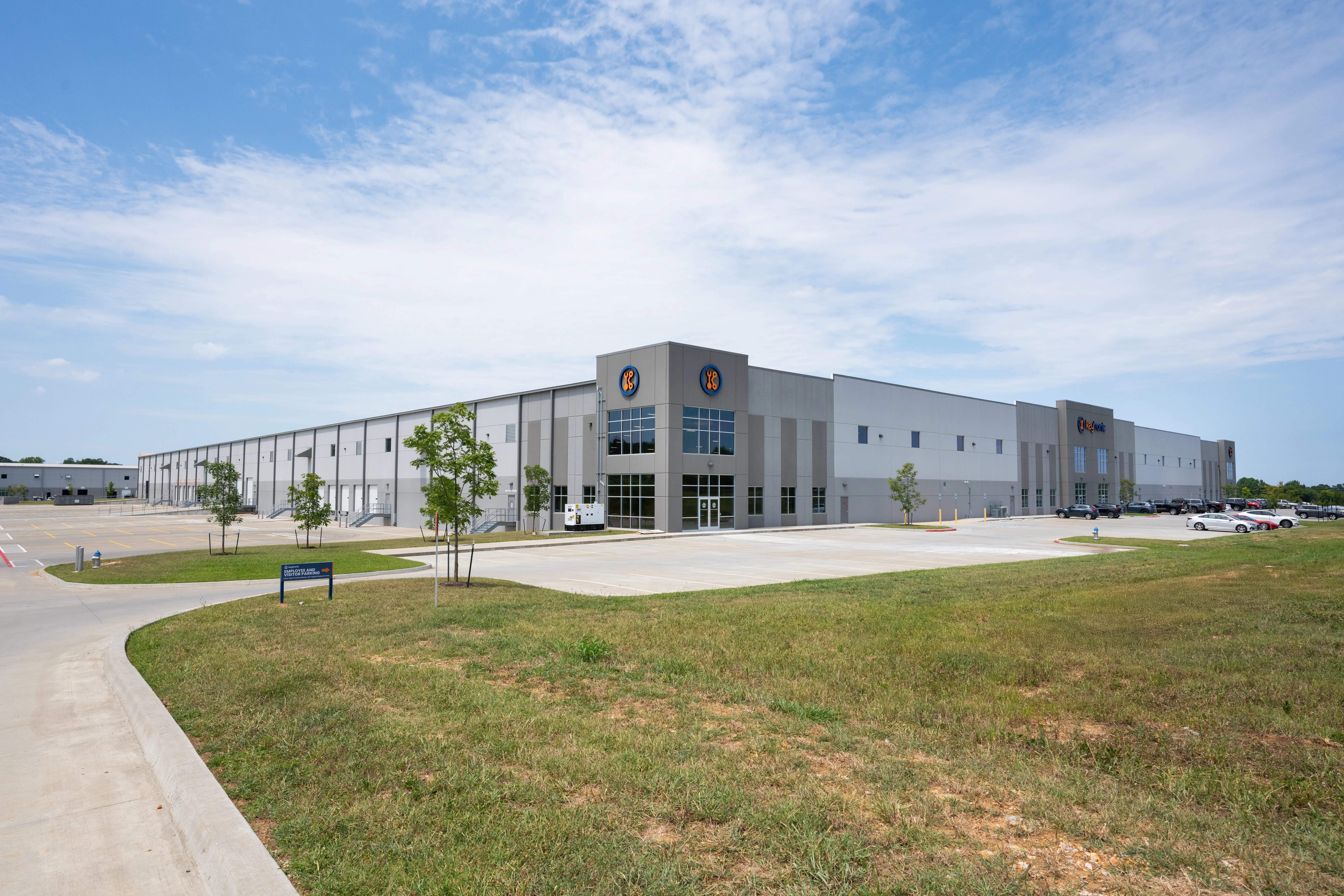 A large, modern industrial warehouse with gray exterior walls, several entrances, and logos on the corners. It is surrounded by a parking lot, a few trees, and open grassy areas under a partly cloudy sky.