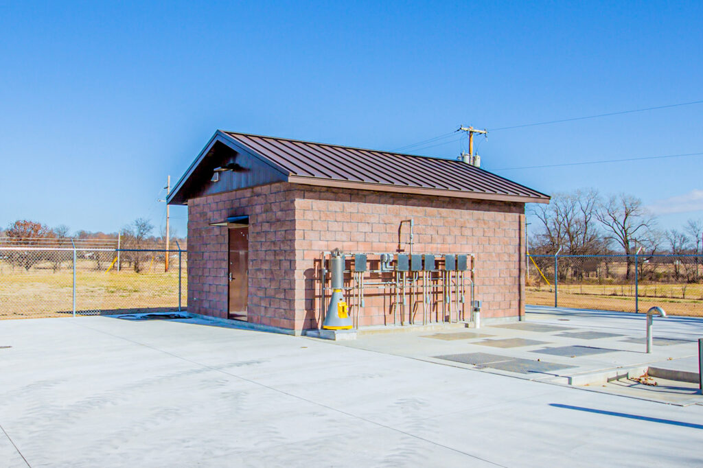 A small brick utility building with a dark roof, outdoor equipment, and meters is situated on a concrete lot, surrounded by a chain-link fence, with grassy fields and leafless trees in the background under a clear blue sky.