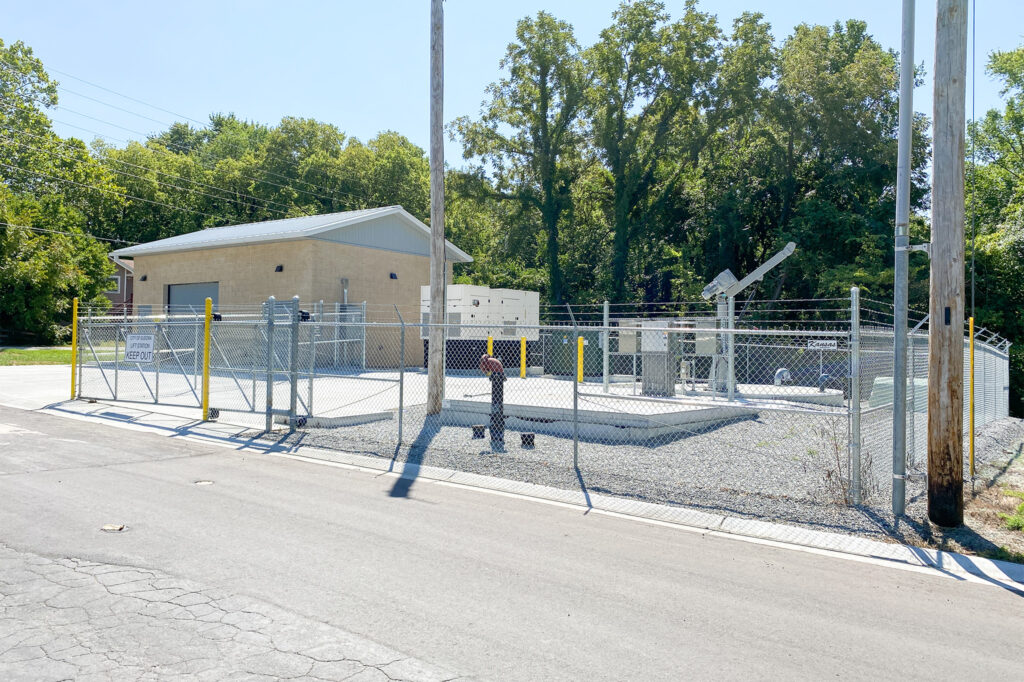 A small, single-story utility building with a fenced area containing electrical equipment and a satellite dish, located by a road with trees in the background on a sunny day.