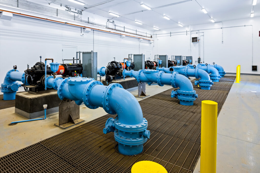 Large blue industrial pipes and pumps are installed in a clean, well-lit room with metal grates on the floor and machinery lined up in rows. Yellow safety posts are also visible.