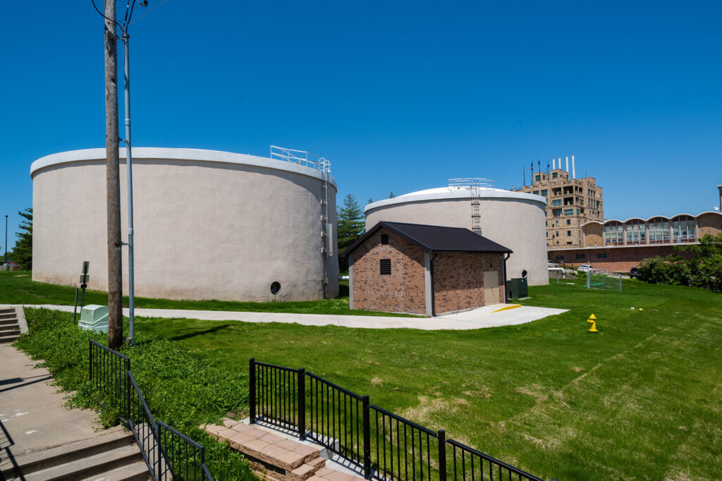 Two large cylindrical storage tanks, a small brick utility building, and a yellow fire hydrant sit on a grassy area under a clear blue sky. A fence and sidewalk appear in the foreground, with more buildings in the background.