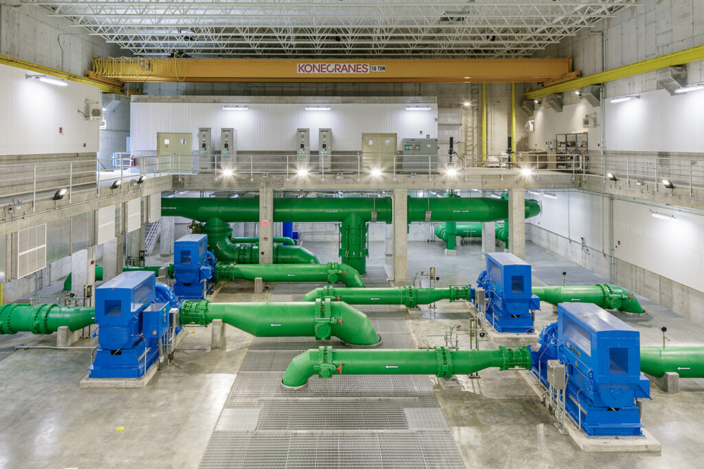 Large industrial facility interior with green pipes, blue industrial machines, concrete floor, and a yellow overhead crane labeled “Konecranes.” Bright lighting and metal railings are visible throughout the space.