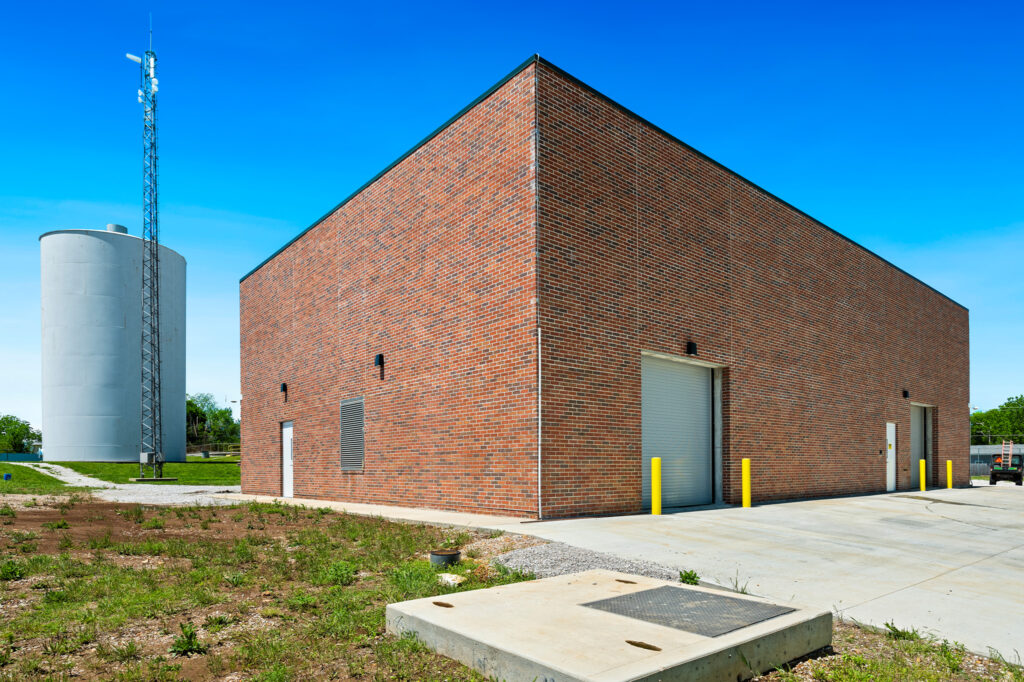 A large brick industrial building with two garage doors, yellow bollards, and sparse vegetation in front. A tall water tower and communication tower stand to the left under a clear blue sky.