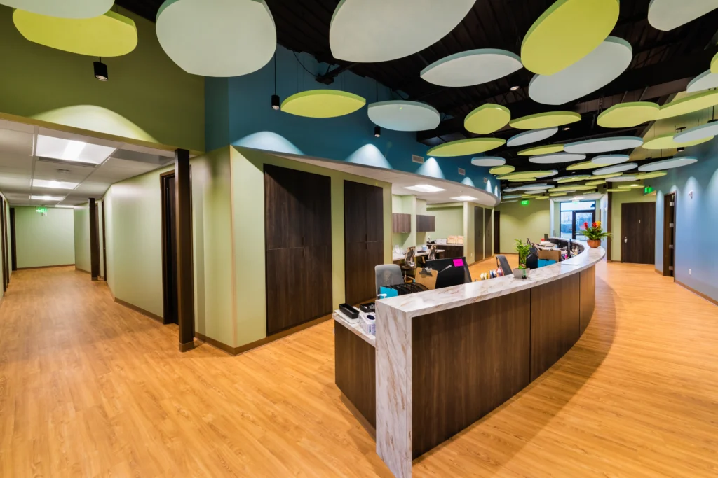 Modern office reception area with a curved desk, light wood flooring, and circular green and white ceiling panels. Hallways and doors lead to other rooms, and the space is brightly lit with contemporary decor.