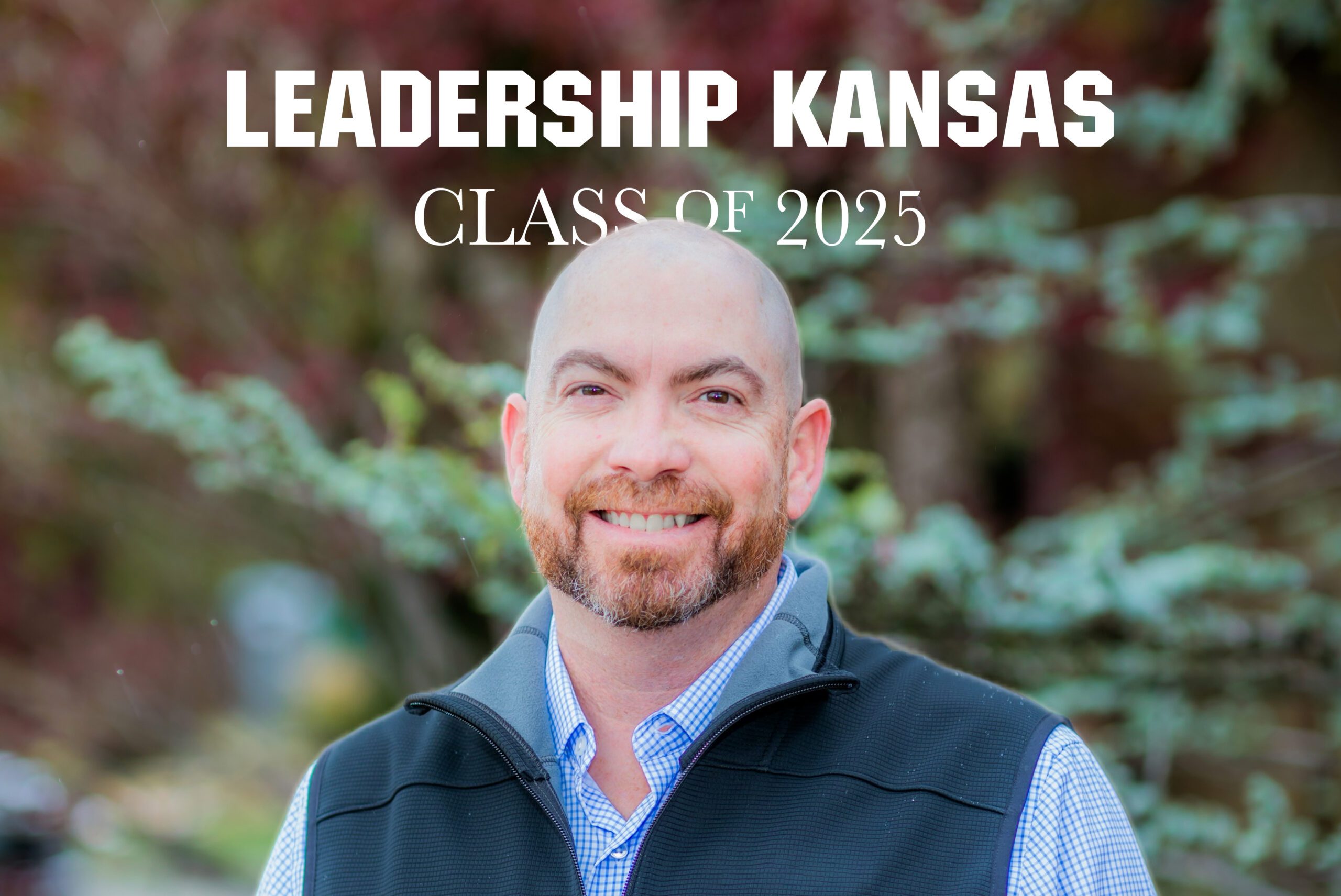 A smiling man with a trimmed beard stands outdoors in front of blurred greenery, wearing a blue checkered shirt and black vest. White text above reads “LEADERSHIP KANSAS CLASS OF 2025.”.