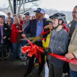 A group of people gather under a pavilion as a man in a suit and two young people wearing bike helmets cut a large red ribbon with oversized scissors, celebrating the opening of a new space or facility.