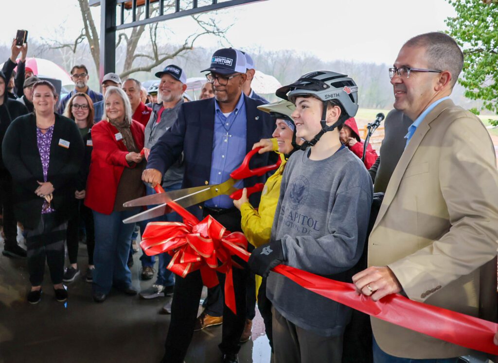 A group of people gathers for a ribbon-cutting ceremony. A smiling boy in a helmet holds a large pair of scissors with two adults, surrounded by onlookers under a covered outdoor area. A red ribbon and bow are in front.