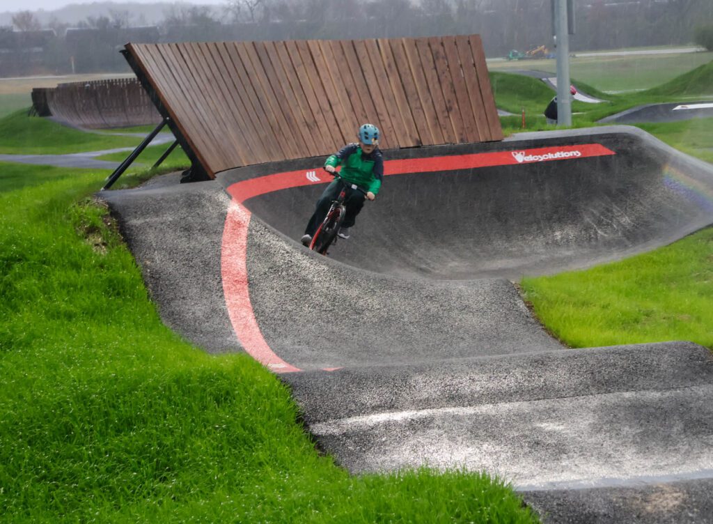 A person wearing a blue helmet and green jacket rides a bike around a curved, paved pump track surrounded by bright green grass on a cloudy day.