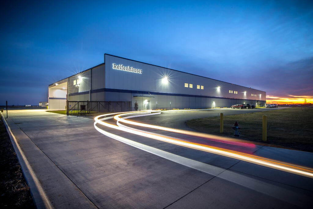 A large industrial building at dusk with “Reifenhäuser” signage, illuminated lights, and a long-exposure effect showing streaks of car lights on the driveway in front.