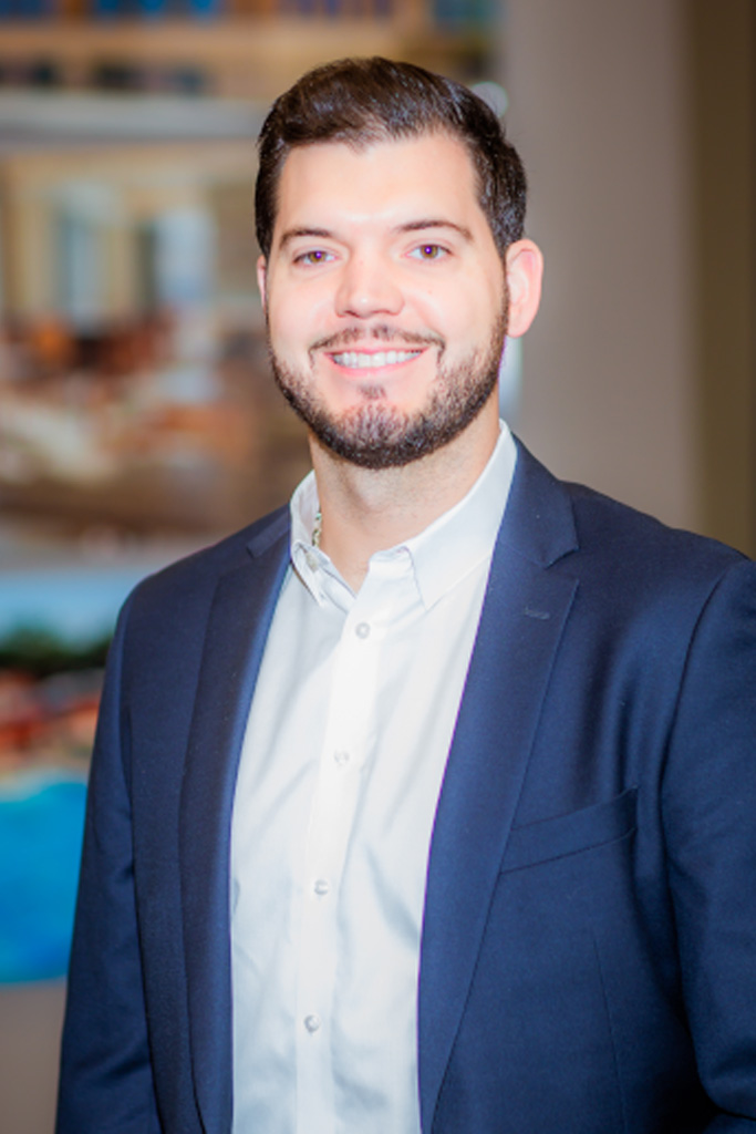 A man with short dark hair and a beard, wearing a navy blazer and white shirt, smiles at the camera. The background is softly blurred, showing abstract colors and shapes.