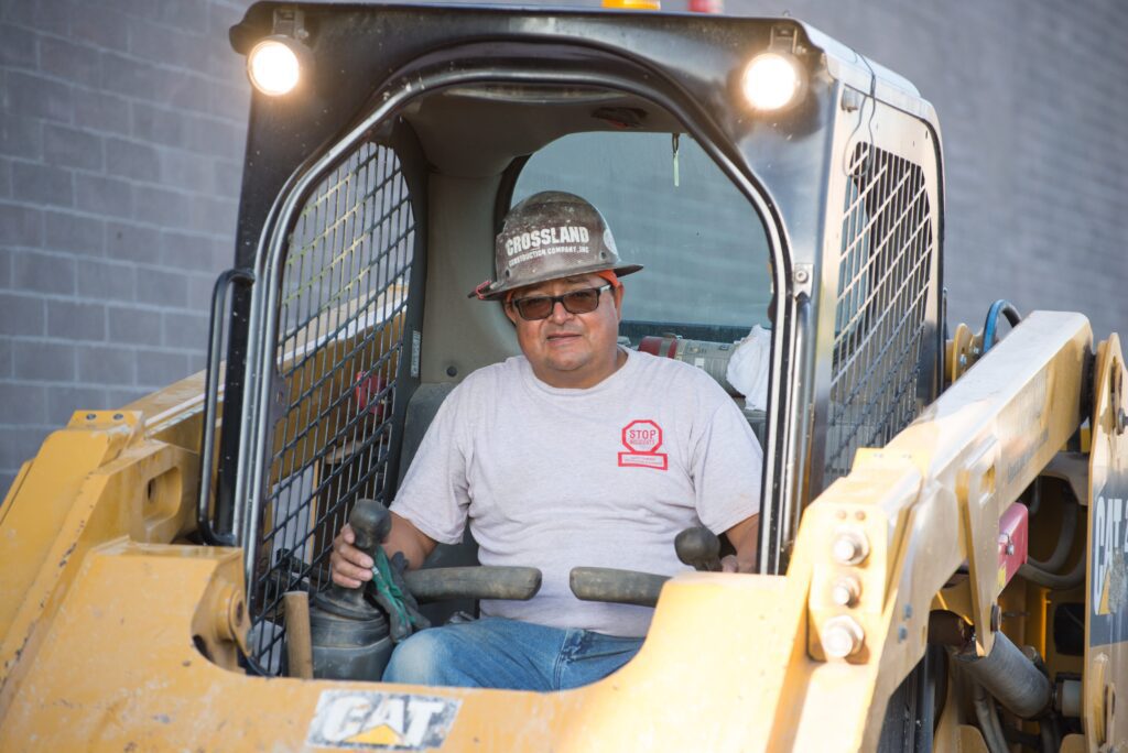 A construction worker wearing safety glasses and a hard hat sits inside the cab of a yellow CAT skid steer loader, operating the controls.