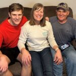 Three people smile while sitting close together on a brown couch. The person on the left wears a red shirt, the person in the middle a light gray shirt, and the person on the right a gray long-sleeve shirt and cap.