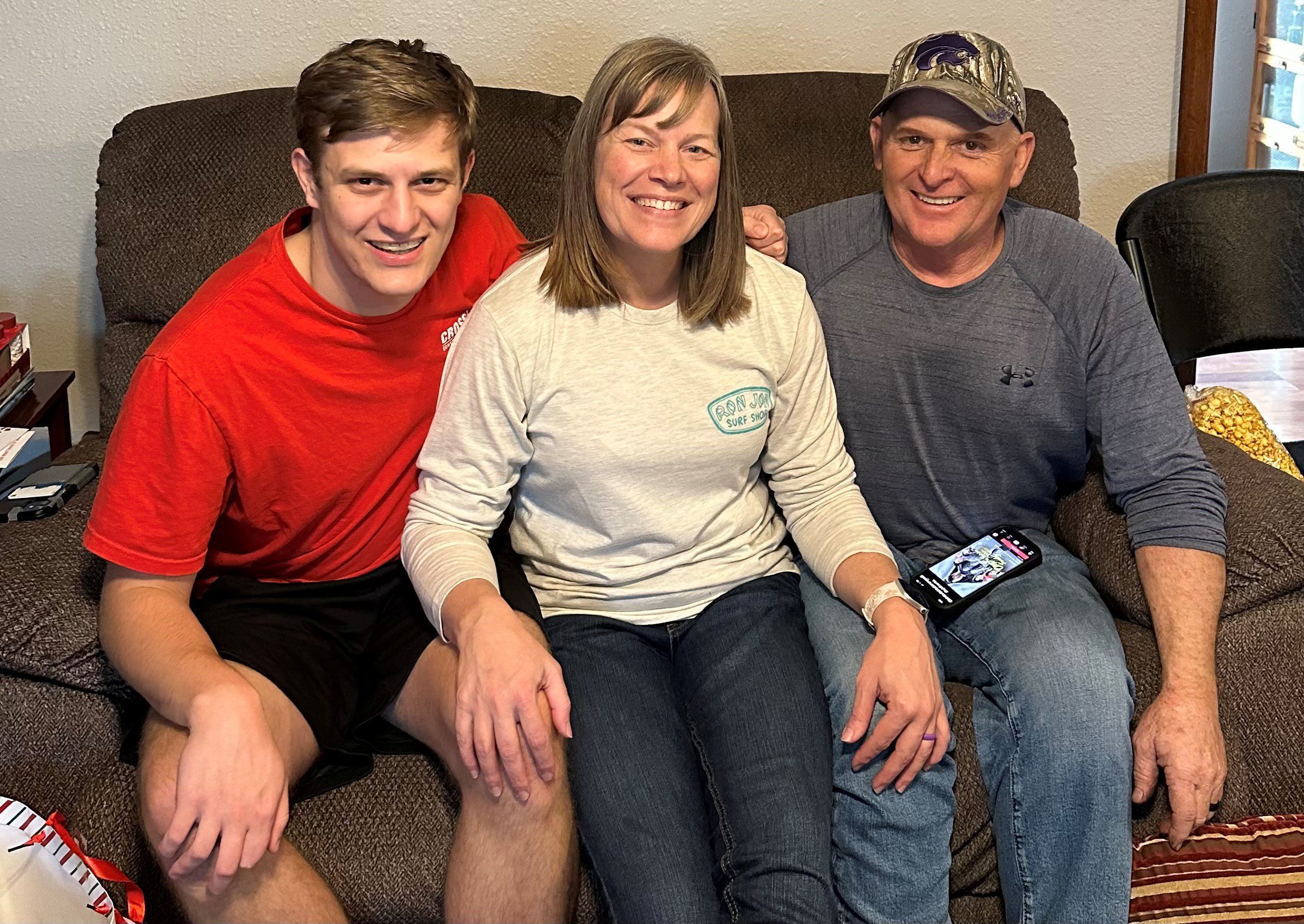 Three people smile while sitting close together on a brown couch. The person on the left wears a red shirt, the person in the middle a light gray shirt, and the person on the right a gray long-sleeve shirt and cap.