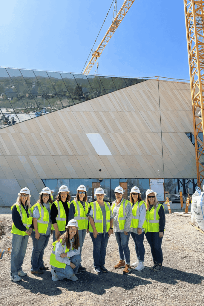 A group of ten people wearing yellow safety vests and hard hats stand in front of a modern building under construction, with a large crane overhead on a sunny day.