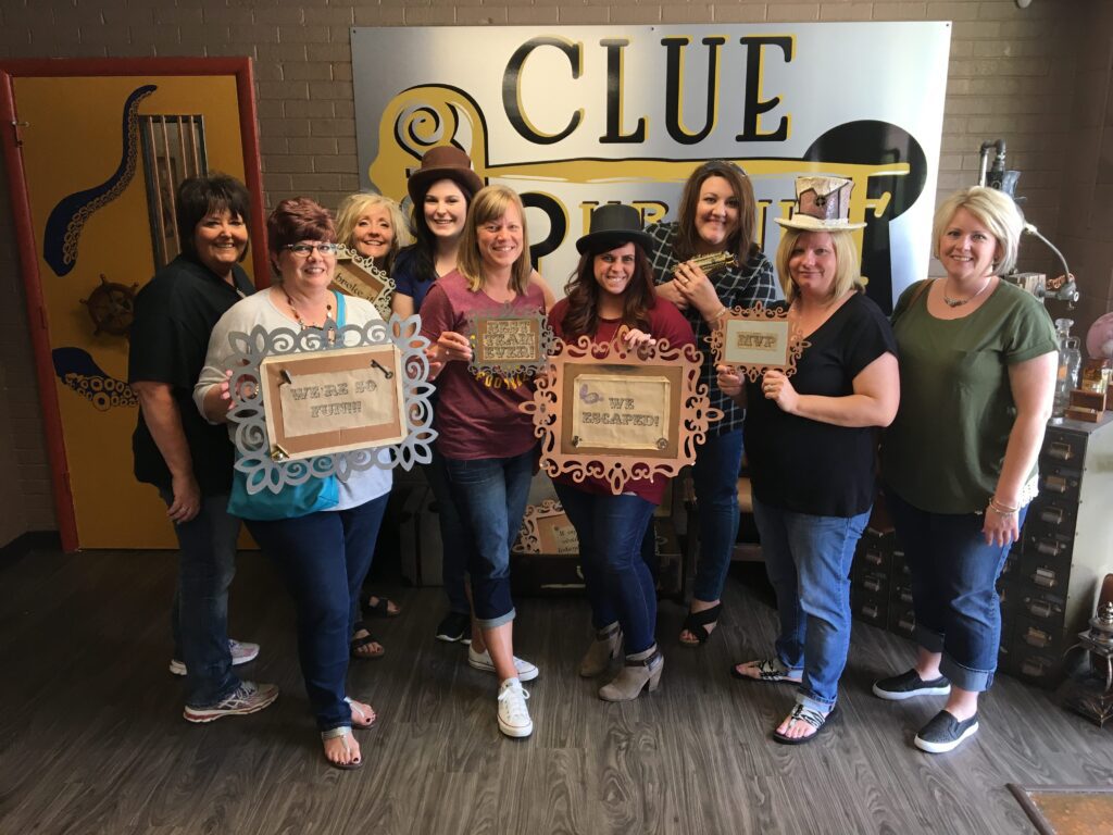 A group of nine women pose and smile in front of a “Clue” sign, holding decorative signs with phrases like “We Escaped” and “Winners” in an escape room setting.