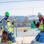 Two construction workers wearing safety gear and masks kneel on a platform, giving peace signs. A highway, cars, and green exit signs are visible in the background under a clear sky.