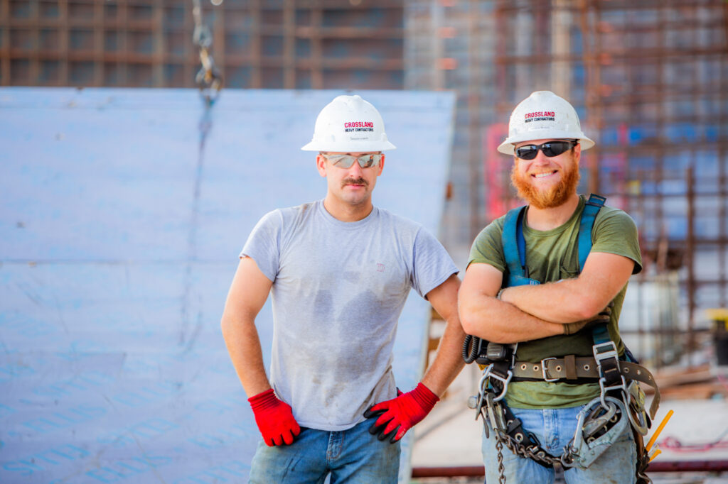 Two construction workers wearing hard hats and safety gear pose and smile at a construction site, with metal structures and equipment visible in the background.