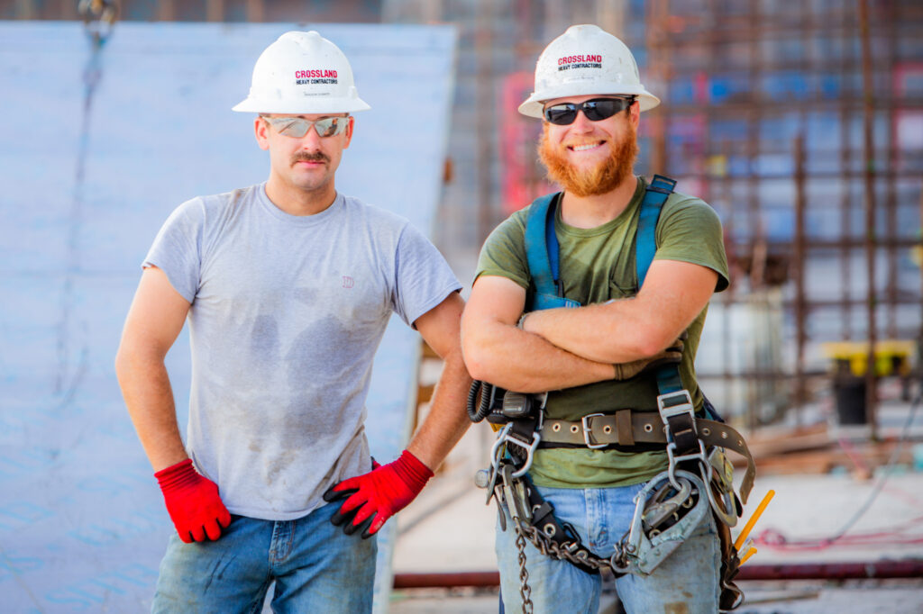 Two construction workers stand together at a job site. Both wear white hard hats, red gloves, and safety gear. One has a beard and sunglasses; the other has a mustache. Scaffolding and construction materials are visible behind them.