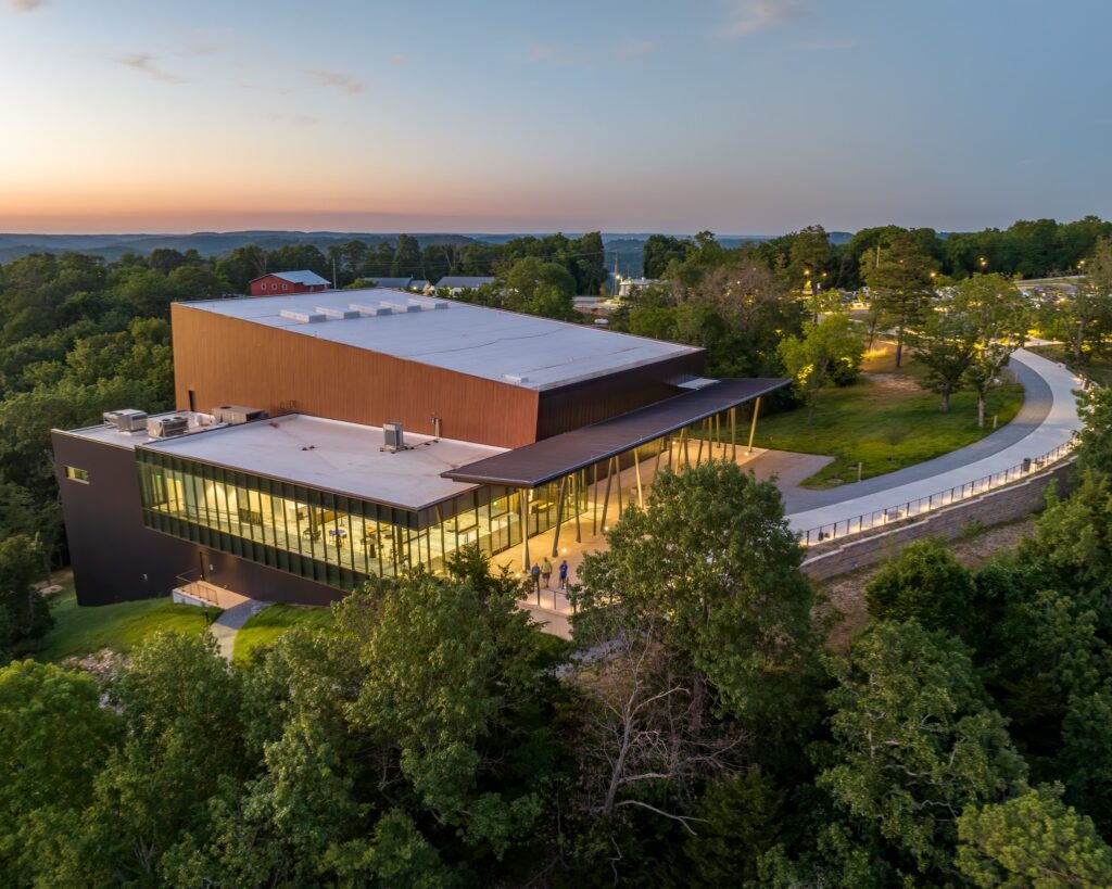 Modern building with large glass windows and a flat roof, surrounded by trees and greenery at sunset. A curved walkway leads to the entrance, and distant hills are visible in the background under a clear sky.