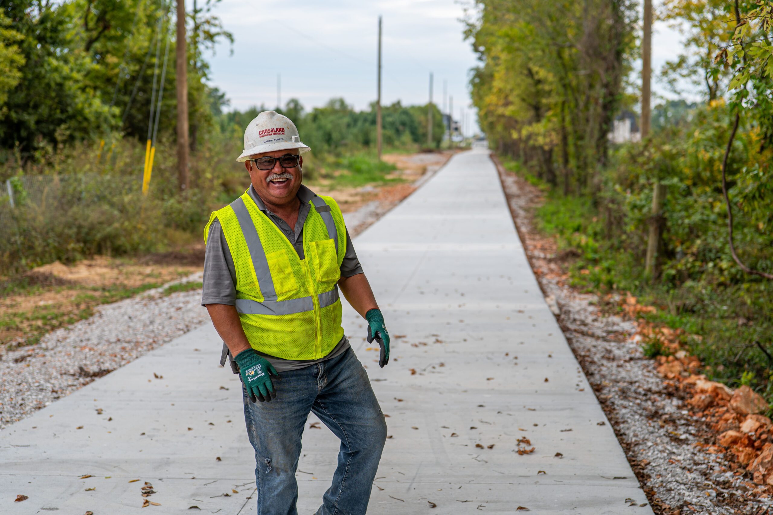 A construction worker in a yellow safety vest and hard hat smiles while standing on a newly paved sidewalk, surrounded by trees and utility poles.