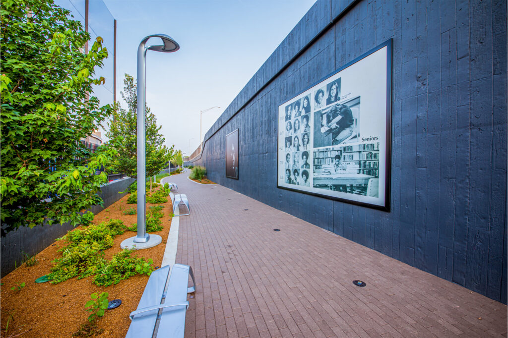 A brick walkway with benches and plants runs alongside a tall dark wall displaying large black-and-white photo collages, under a clear sky.
