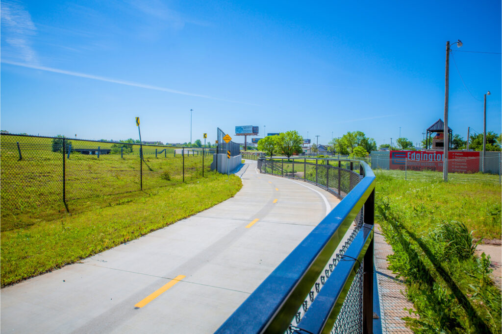 A paved biking and walking trail curves through a grassy area, bordered by chain-link fences, under a clear blue sky with billboards and a playground visible in the background.