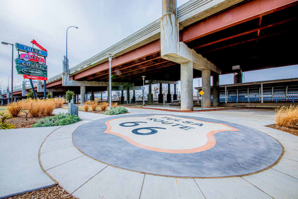 A large Tulsa Route 66 emblem is painted on the sidewalk under a highway overpass, with the Tulsa Auto Court sign and landscaping visible nearby.
