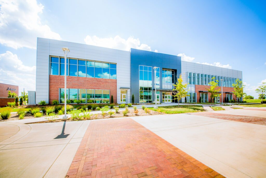 A modern two-story building with large windows, brick and metal exterior, surrounded by landscaped greenery and a wide paved walkway under a bright blue sky with scattered clouds.