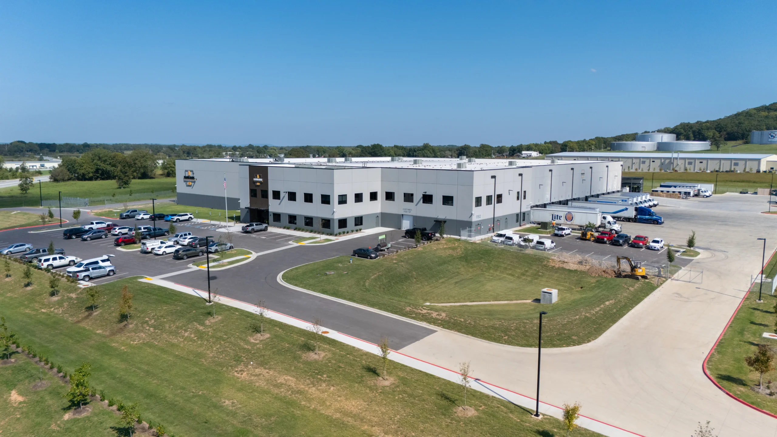 Aerial view of a large, modern UPS warehouse facility with parked cars and delivery trucks outside, surrounded by green lawns and trees under a clear blue sky.