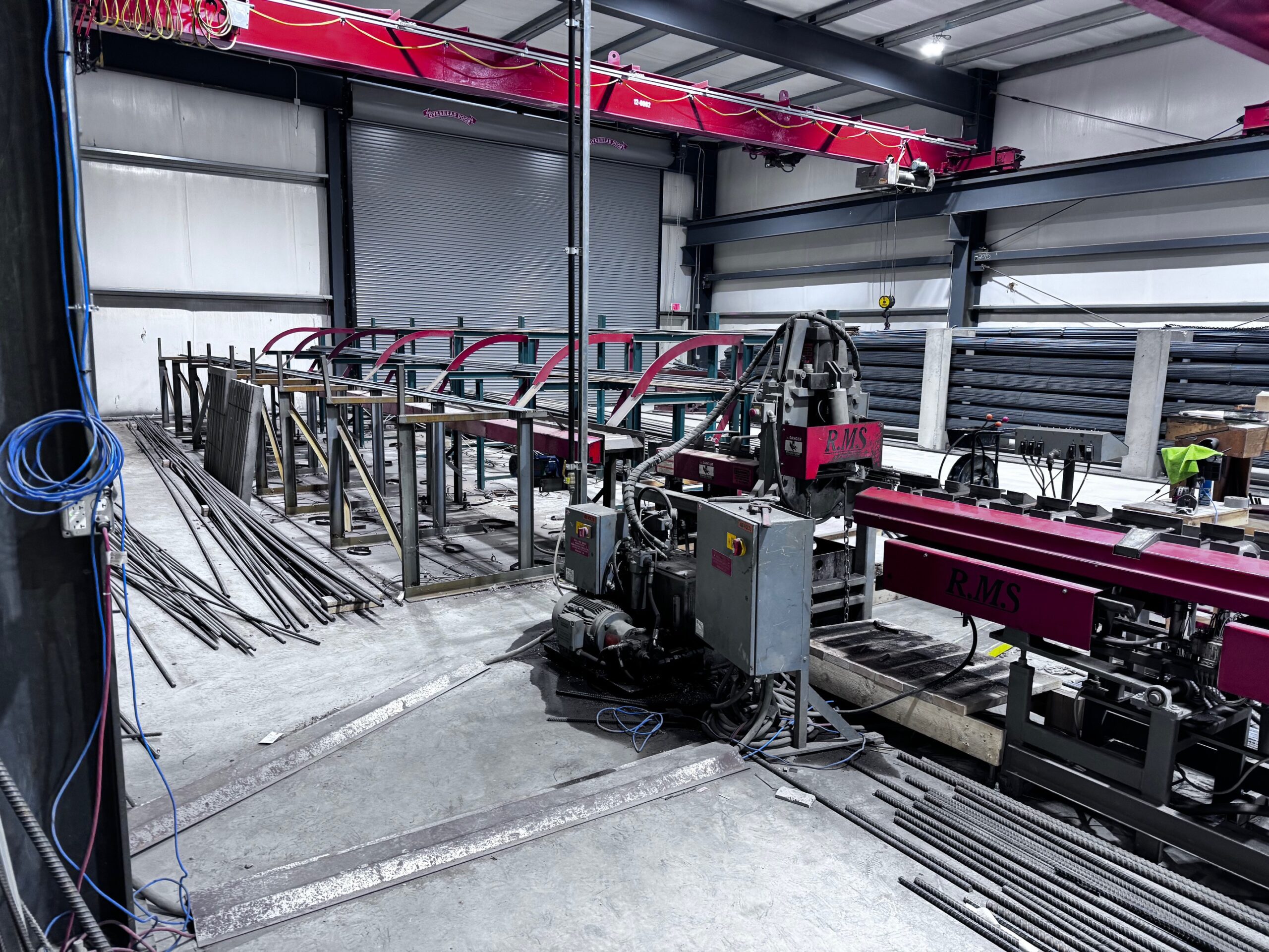 Metal fabrication workshop with industrial machinery, including a red RMS saw, metal bars, and partially assembled structures. The area is spacious with concrete floors and exposed metal beams.