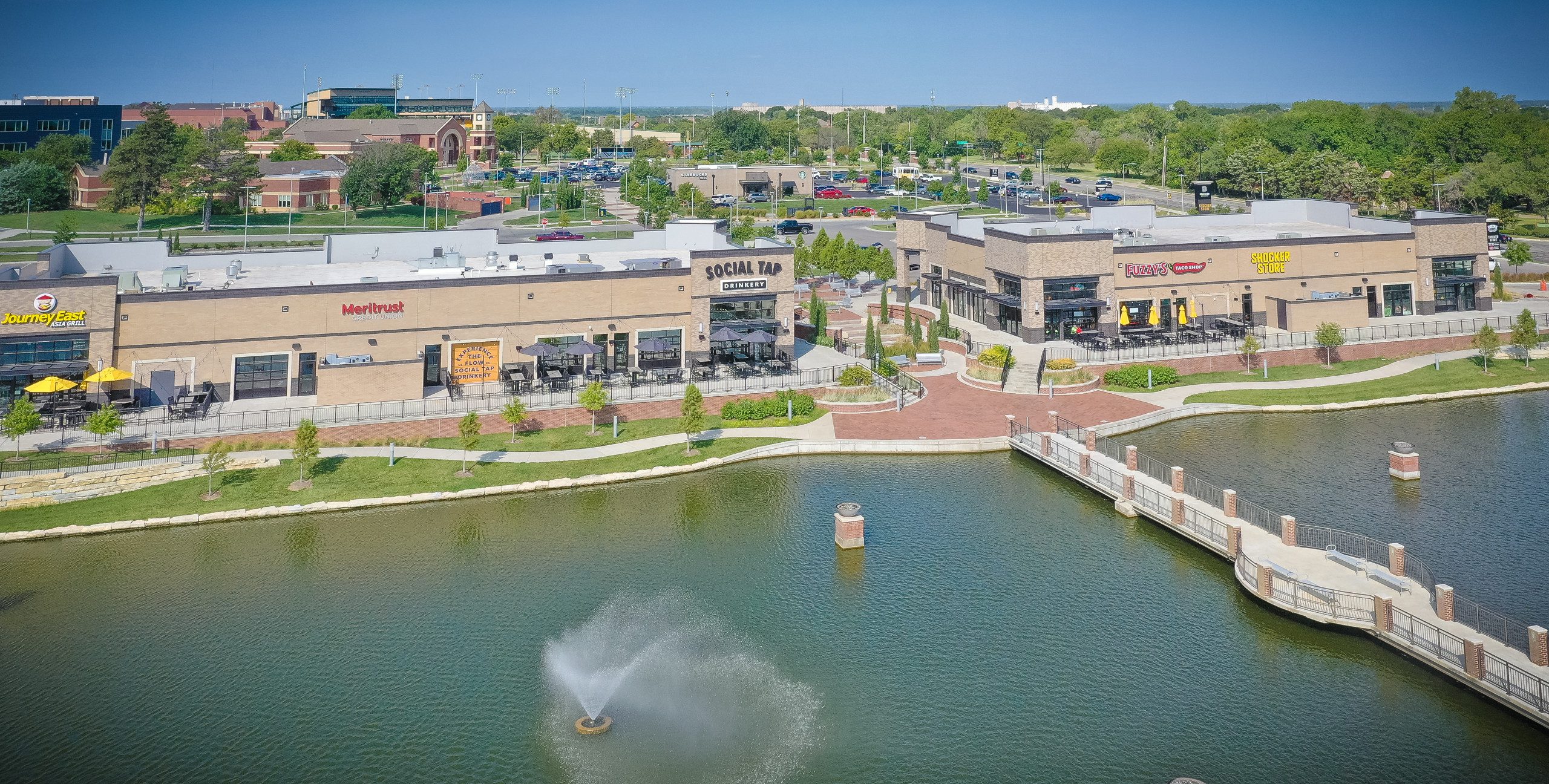 A shopping plaza with several restaurants and shops overlooks a pond with a water fountain. Outdoor seating areas, landscaped paths, and parking lots are visible, with green trees and grass throughout.