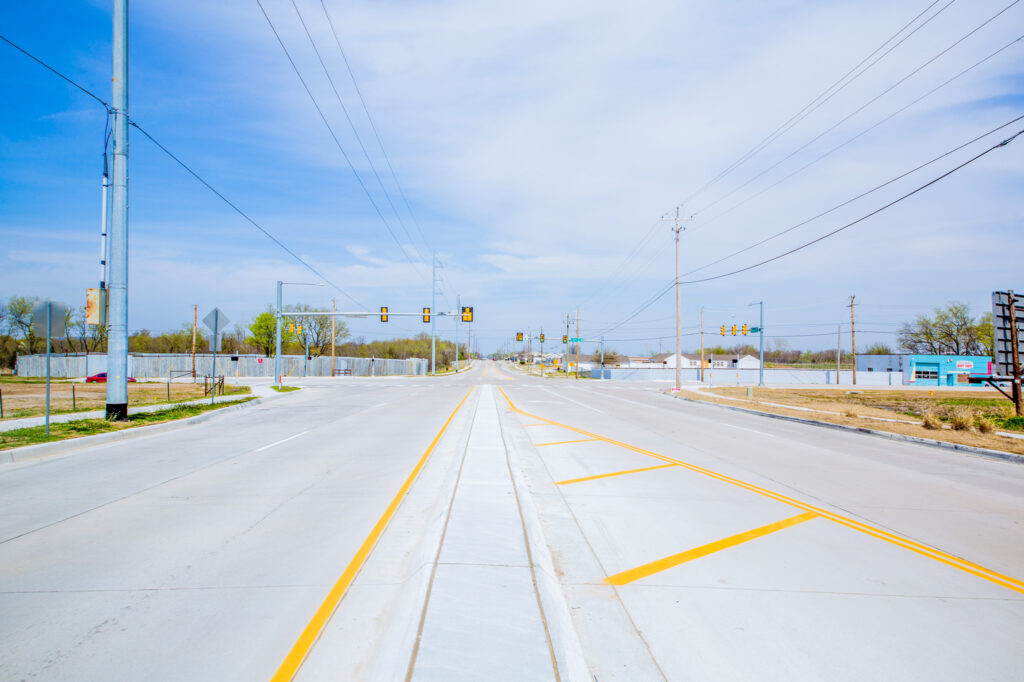 A wide, empty intersection with traffic lights under a clear blue sky. Power lines and utility poles line both sides of the road, and some buildings and trees are visible in the background.
