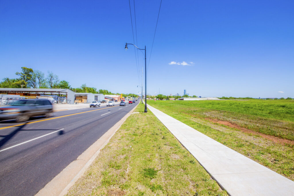 A wide road with moving cars runs beside a long sidewalk and green grassy field under a clear blue sky. Buildings and trees line the left side, while an open field is on the right.