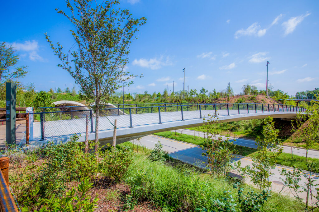 A modern pedestrian bridge with metal railings spans over a road, surrounded by lush greenery and young trees under a sunny blue sky.