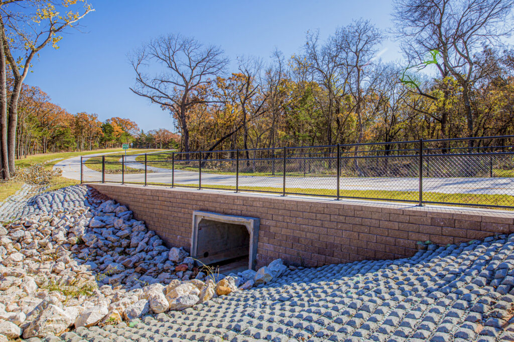 A paved path with a metal fence crosses over a small culvert surrounded by rocks and trees with autumn leaves under a clear blue sky.
