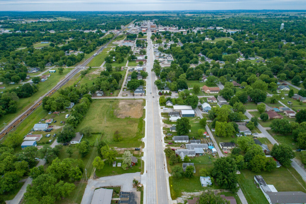 Aerial view of a small town with a straight road running through the center, houses and trees on both sides, and green fields in the background under a cloudy sky.