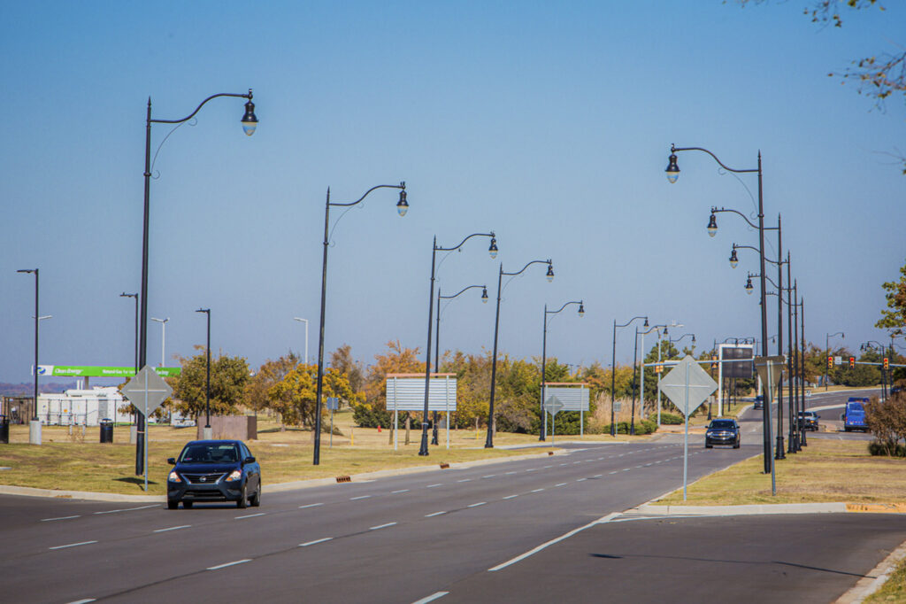 A wide, empty street with multiple streetlights, a few cars driving, green highway signs in the distance, and trees lining both sides under a clear blue sky.
