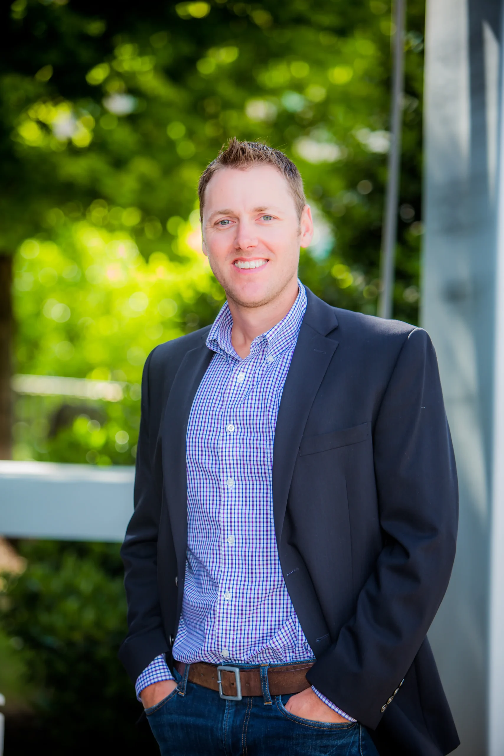 A man wearing a dark blazer and checked shirt smiles while standing outdoors with greenery and sunlight in the background.