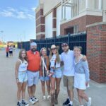 Six people pose together outside a brick building on a sunny day. Most are wearing casual, sporty outfits, with some in orange and white colors. The group smiles at the camera, standing on a paved walkway near a black metal fence.