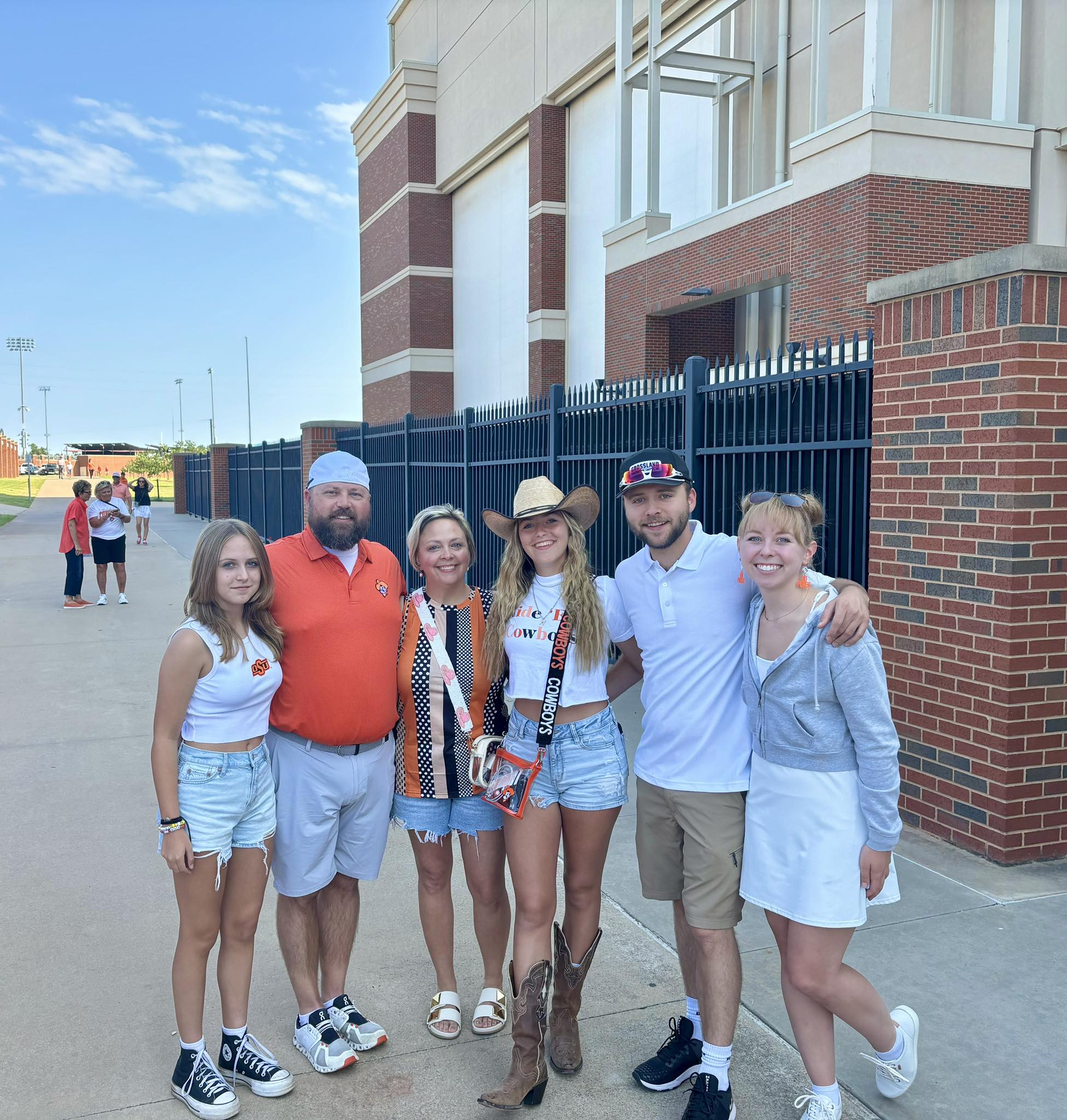 Six people pose together outside a brick building on a sunny day. Most are wearing casual, sporty outfits, with some in orange and white colors. The group smiles at the camera, standing on a paved walkway near a black metal fence.