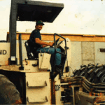 A man wearing a cap and jeans sits and smiles on the seat of a large construction vehicle in an industrial area, with tires and a concrete wall visible in the background.