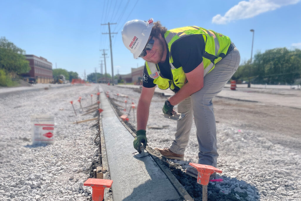A construction worker in a safety vest and helmet smooths wet concrete on a road under construction, with traffic cones and buildings visible in the background.